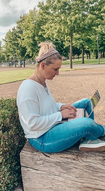 Tatjana Fadda, eine Virtuelle Assistentin und Expertin für E-Mail-Marketing-Service, sitzt auf einem Baumstamm in einem Park und benutzt ihren Laptop.