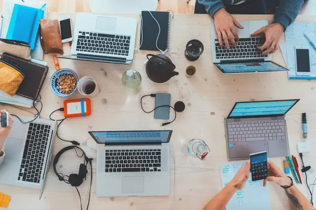 Draufsicht auf einen Arbeitsplatz mit mehreren Laptops, Telefonen, Notizbüchern, Kopfhörern und einer Schüssel Snacks auf einem Tisch.