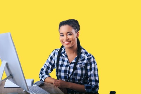 Eine junge Frau sitzt an einem Schreibtisch mit einem Computer vor gelbem Hintergrund.