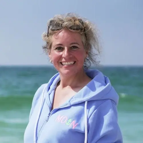 Eine Frau mit lockigem blondem Haar, die einen hellblauen Kapuzenpulli und eine Sonnenbrille auf dem Kopf trägt, steht lächelnd vor dem Meer unter einem klaren Himmel.