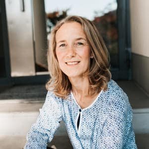 Eine Frau mit schulterlangen blonden Haaren, die ein blau-weiß gemustertes Oberteil trägt, sitzt auf einer Steintreppe.