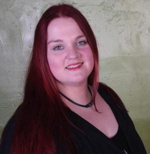 Eine Person mit langen roten Haaren, die ein schwarzes Shirt und eine schmale Halskette trägt.