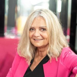Eine lächelnde Frau mit schulterlangen blonden Haaren, die ein schwarzes Oberteil und einen leuchtend rosa Blazer trägt.