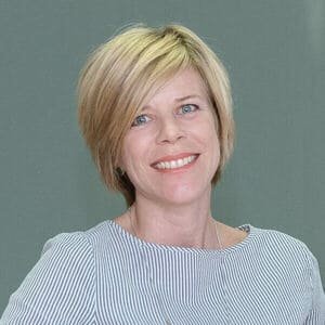 Porträtfoto einer lächelnden Frau mit schulterlangen blonden Haaren, die ein blau-weiß gestreiftes Shirt trägt.
